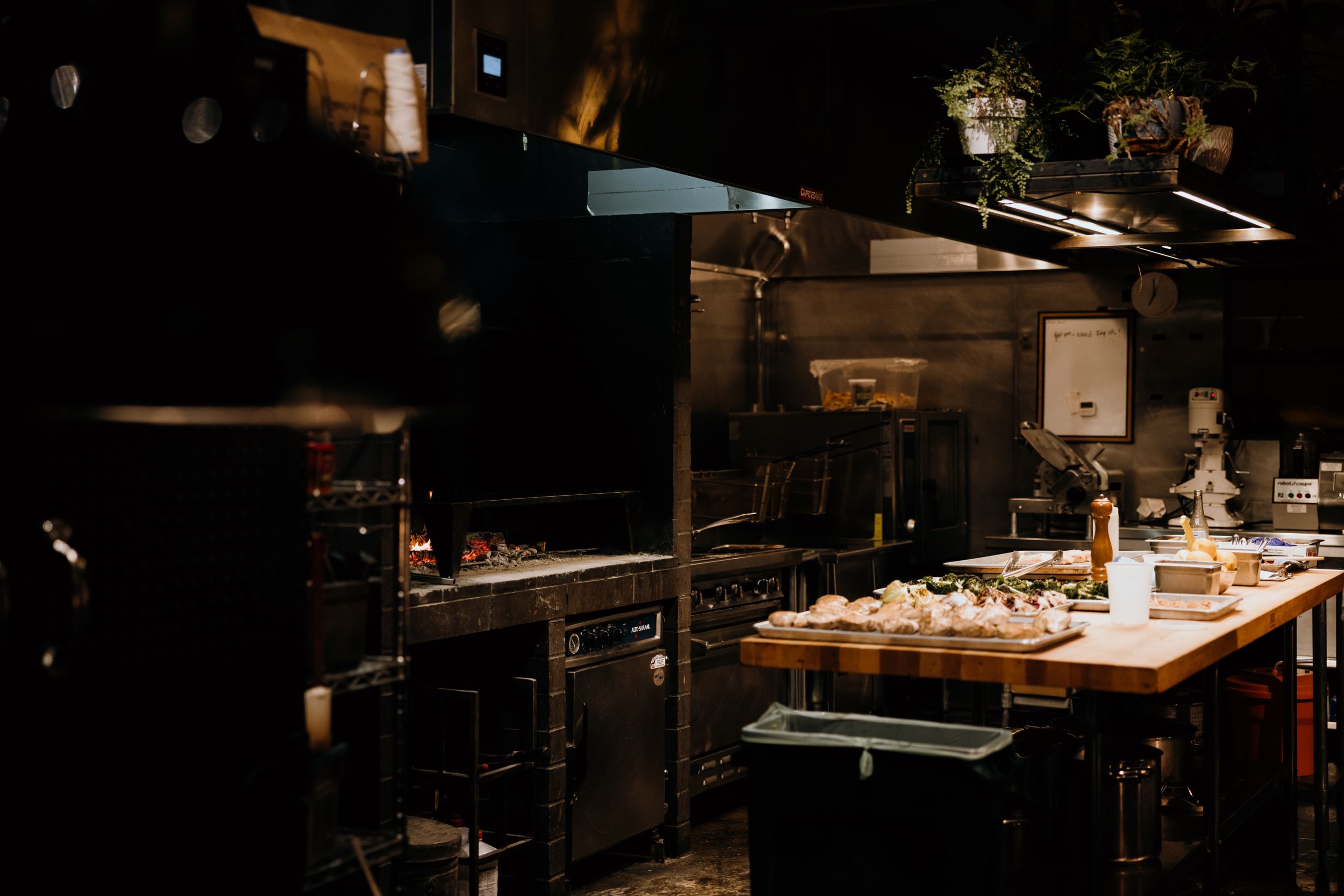 A working kitchen at low light — live fire on the grill, sheet trays of prepped food waiting on the butcher-block pass.