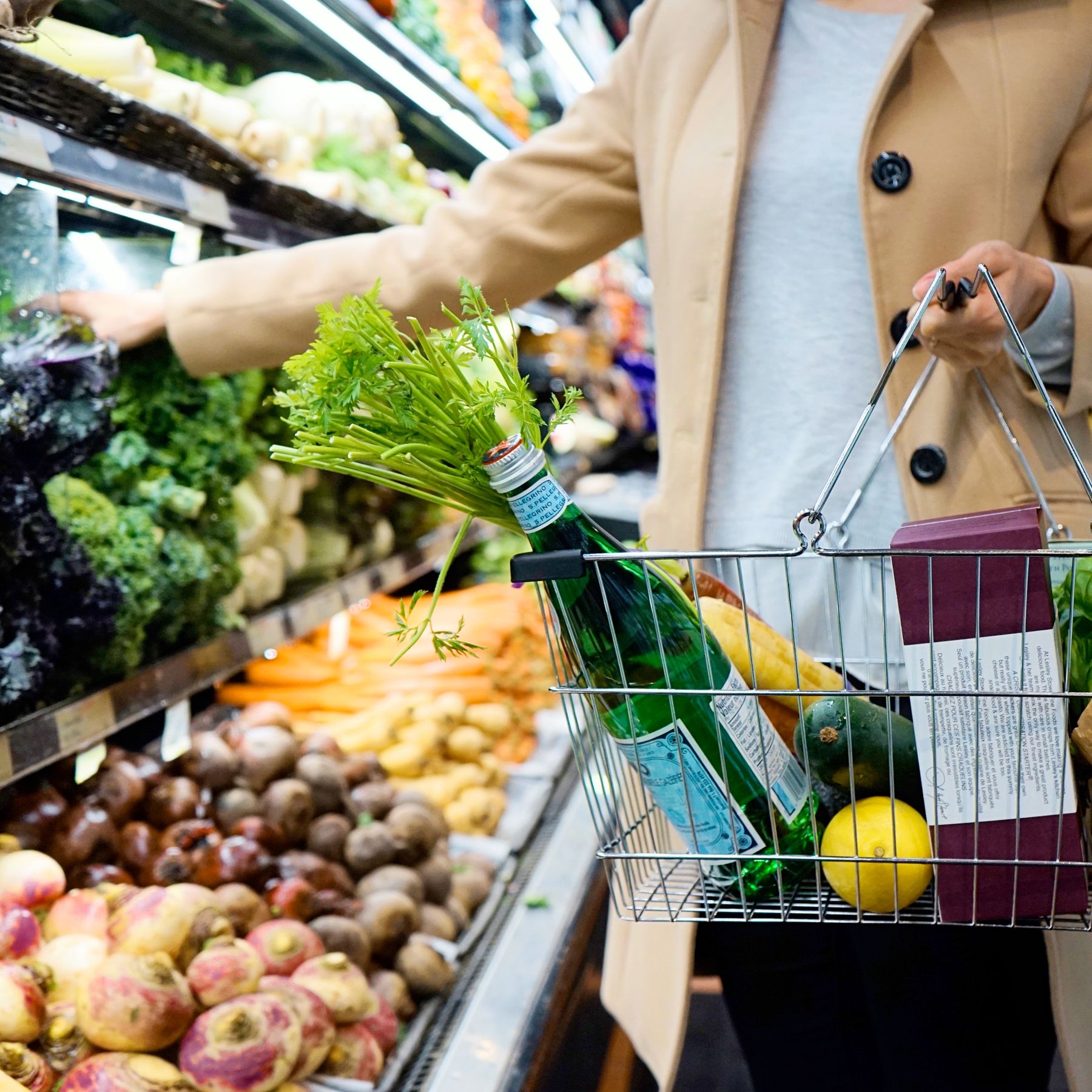 A shopper's hand hovering over a grocery shelf, basket of fresh greens beside her — the moment before flipping the bottle over.
